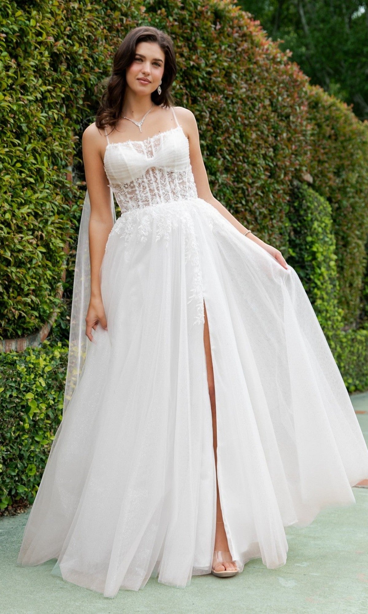 A woman standing outdoors in front of a tall green hedge wears the Cinderella Couture 4217 long formal dress, featuring a white, sleeveless design with a pearl-beaded lace bodice and slit skirt.